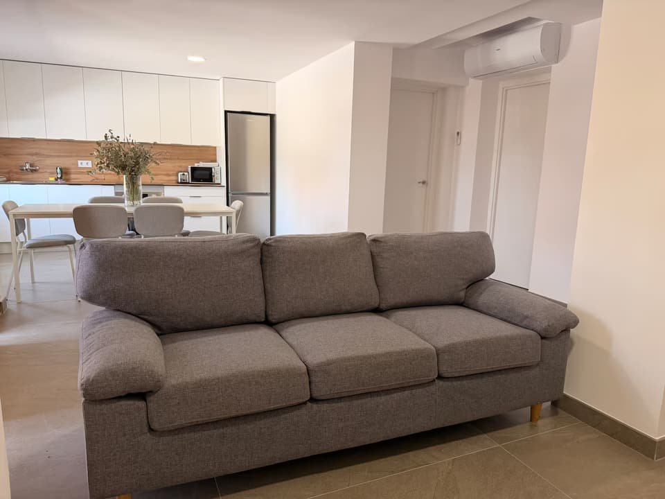 Salón de la Casa A con sofá gris de tres plazas y, al fondo, la cocina y el comedor