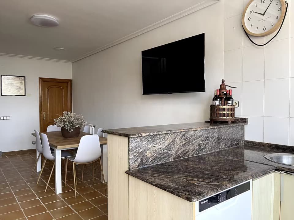 Barra de cocina de la Casa B con encimera de granito, televisión y comedor al fondo
